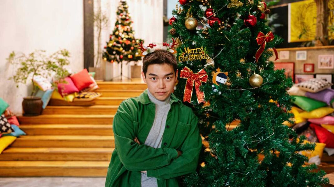 Hombre joven con brazos cruzados y diadema de renos posa junto a un árbol de Navidad decorado con lazos rojos, en un interior con escalones de madera y cojines.