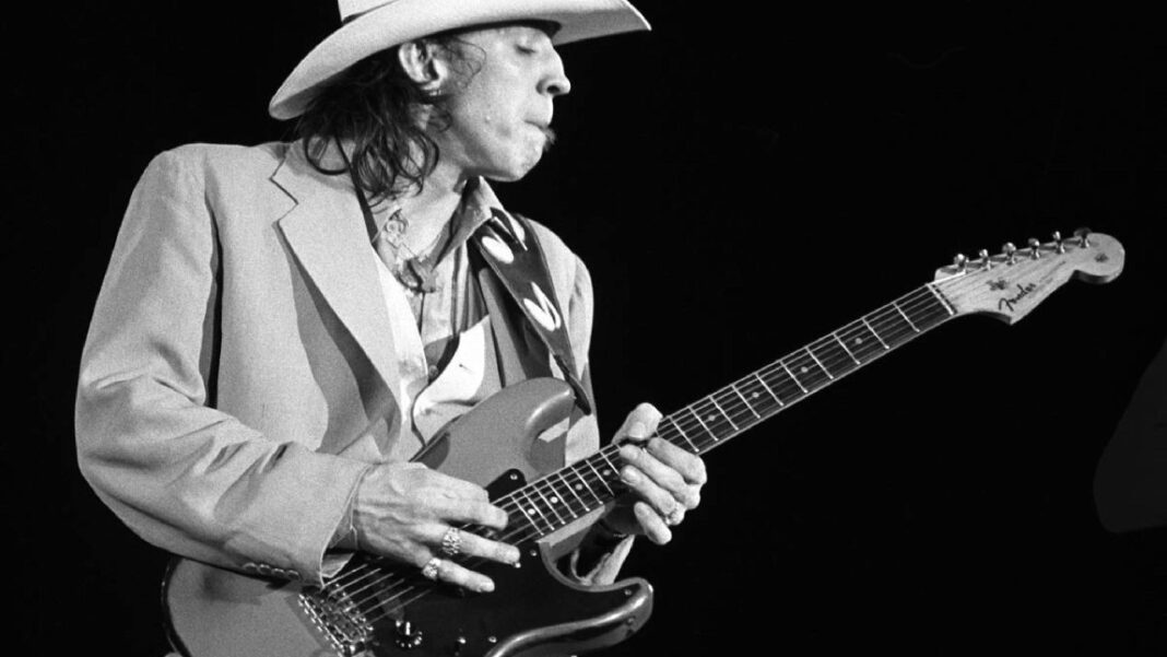 Fotografía en blanco y negro de un músico con sombrero tejano y americana clara tocando una guitarra eléctrica, de perfil sobre fondo negro.