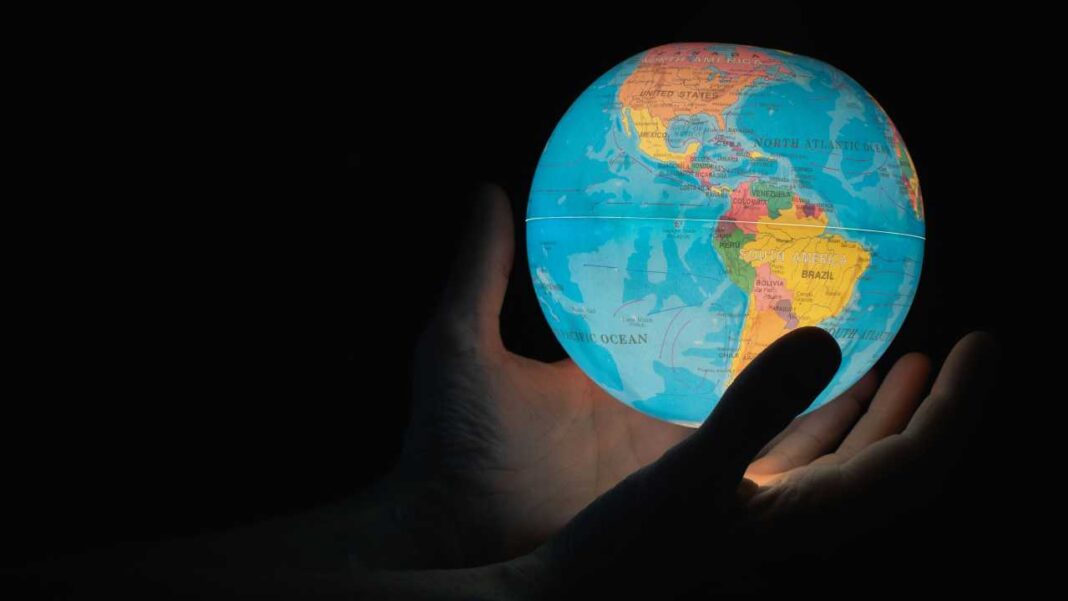Una mano sostiene un globo terráqueo iluminado en la oscuridad; se ven océanos y continentes como América del Norte y del Sur.