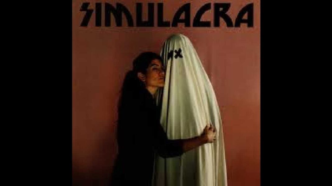Una mujer de cabello oscuro y ropa negra abraza a una figura cubierta con una tela clara, frente a un fondo marrón rojizo con el texto SIMULACRA.