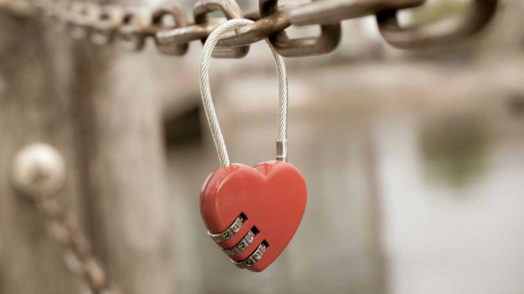 Candado con forma de corazón rojo colgado de una cadena metálica, con ruedas numéricas visibles y un fondo desenfocado.