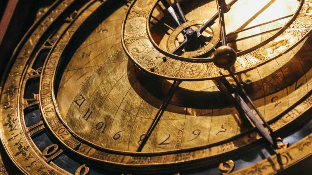 Primer plano de un reloj astronómico dorado con anillos concéntricos, números romanos y arábigos, agujas metálicas y superficies grabadas bajo luz cálida.
