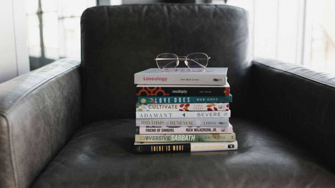 Una pila de nueve libros con títulos visibles descansa sobre un sillón de cuero oscuro, y unas gafas de montura metálica están encima.