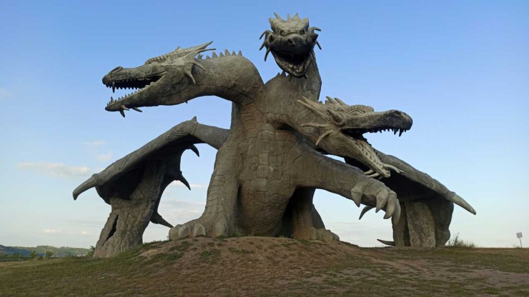 Escultura monumental de un dragón de tres cabezas con alas extendidas sobre un montículo de tierra bajo un cielo azul.