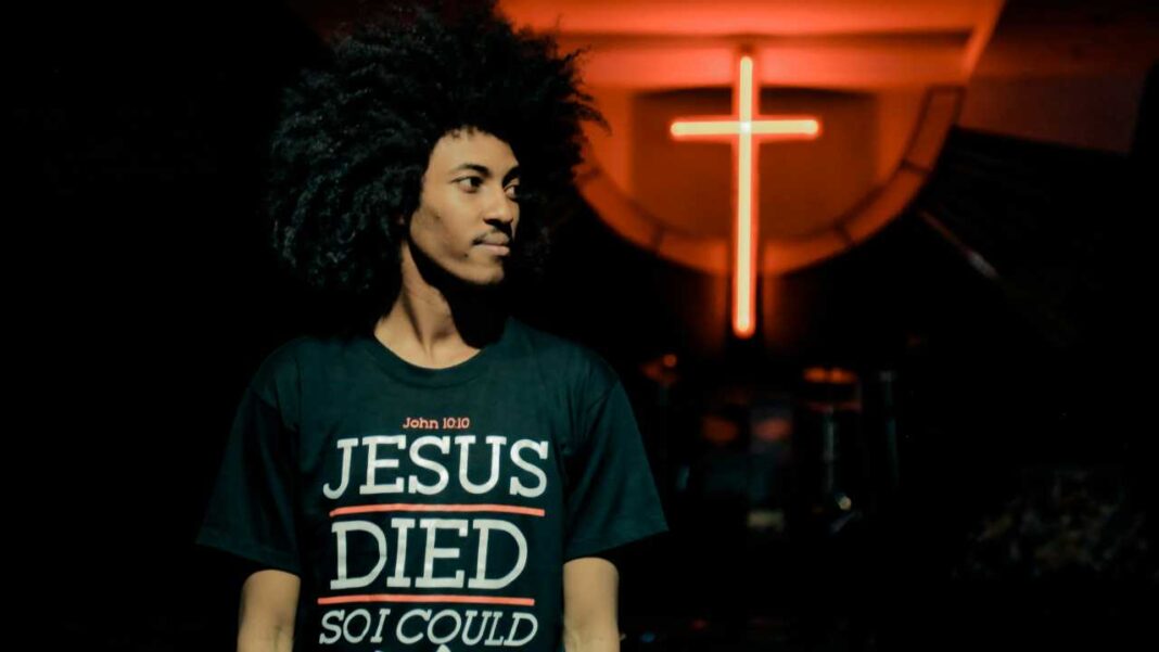Un joven de cabello afro y camiseta verde oscura con texto religioso mira de lado frente a una cruz roja iluminada en un fondo oscuro.