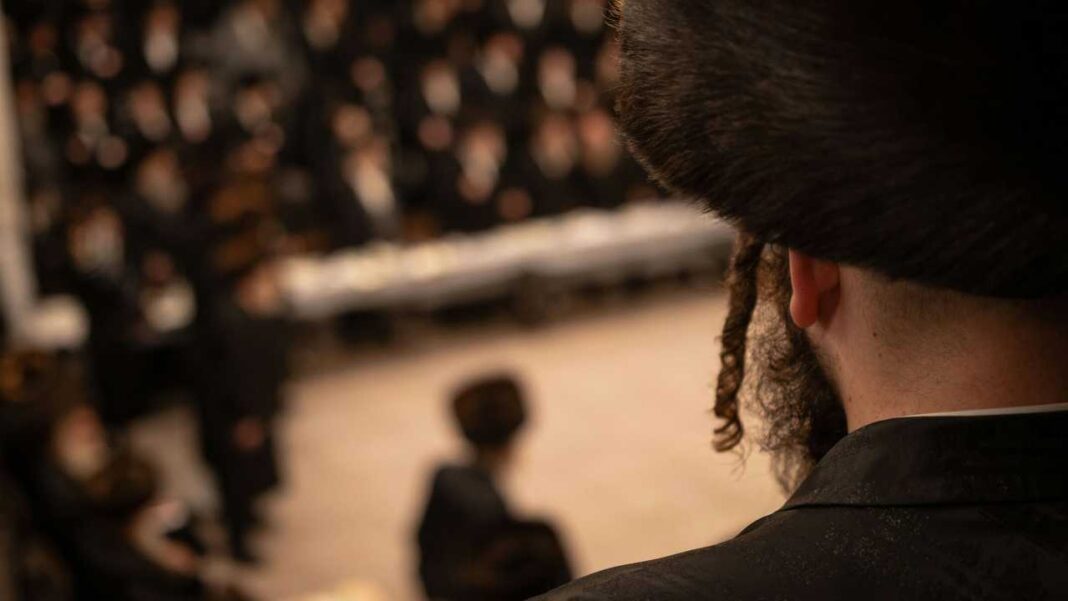 Primer plano de la nuca y el hombro de un hombre con cabello oscuro y rizo lateral, ante una multitud desenfocada en un interior.