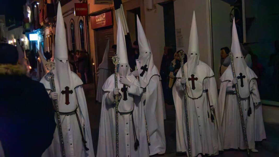 Varias personas con túnicas blancas y capirotes puntiagudos caminan de noche por una calle estrecha, llevando bastones y cruces rojas en el pecho.