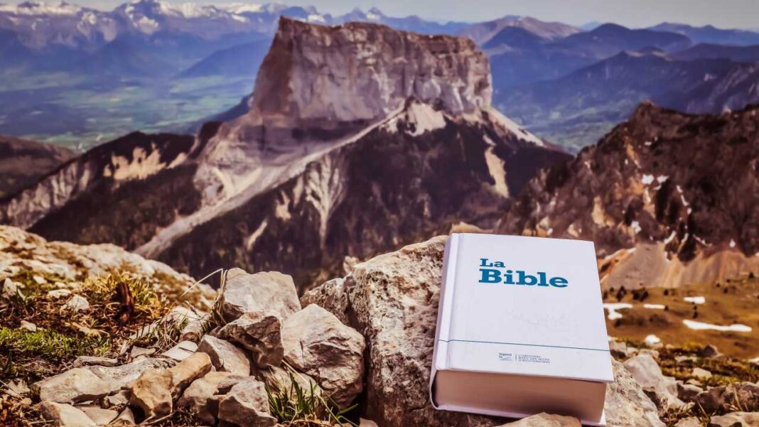 Un libro blanco con el texto La Bible descansa sobre piedras en primer plano, con grandes montañas rocosas y un valle al fondo.