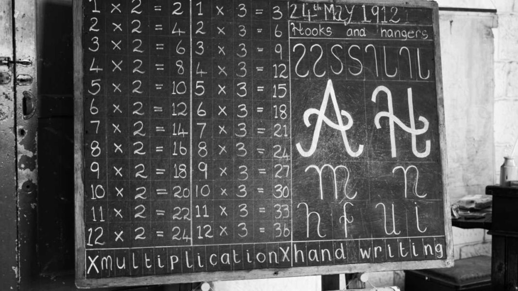 Pizarra escolar en blanco y negro con tablas de multiplicar del 2 y del 3, la fecha 24 de mayo de 1912 y ejemplos de caligrafía.