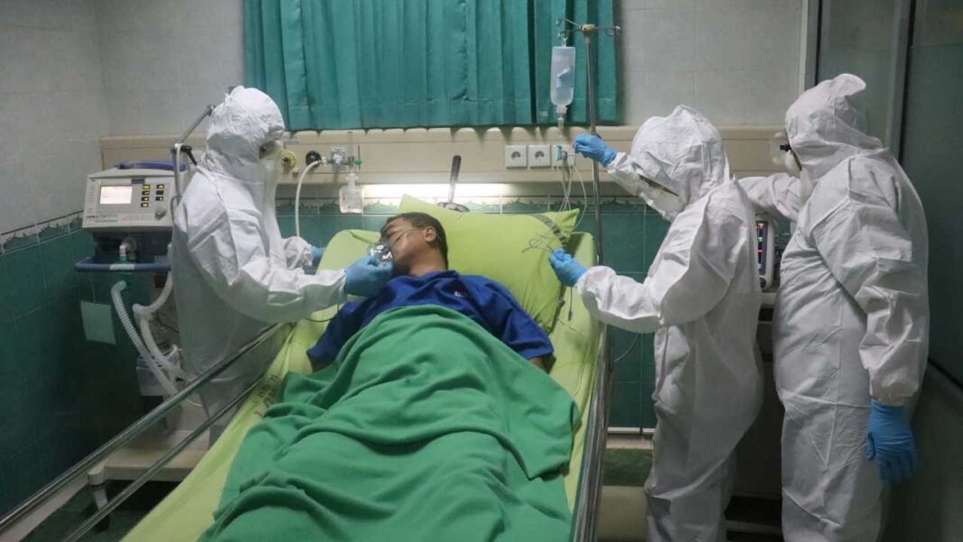 Paciente acostado en cama de hospital rodeado por tres personas con trajes de protección blancos y guantes azules; equipo médico y suero al fondo.