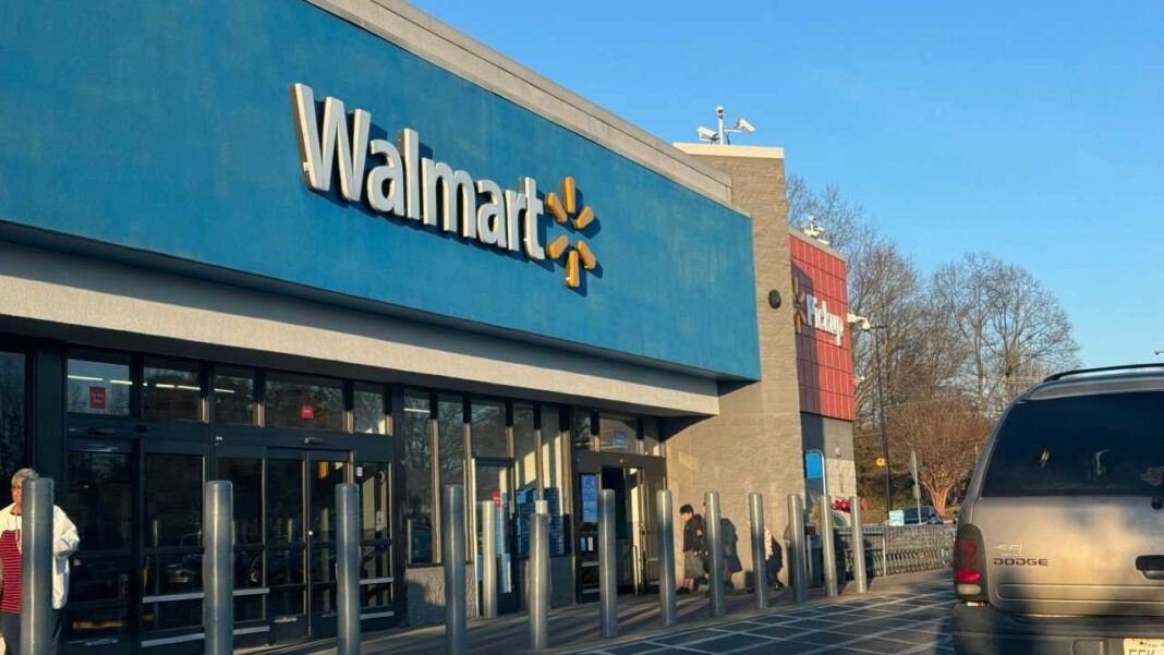 Fachada azul de Walmart con letras blancas y logo amarillo, puertas de vidrio, bolardos, una persona junto a la entrada y una camioneta gris a la derecha.