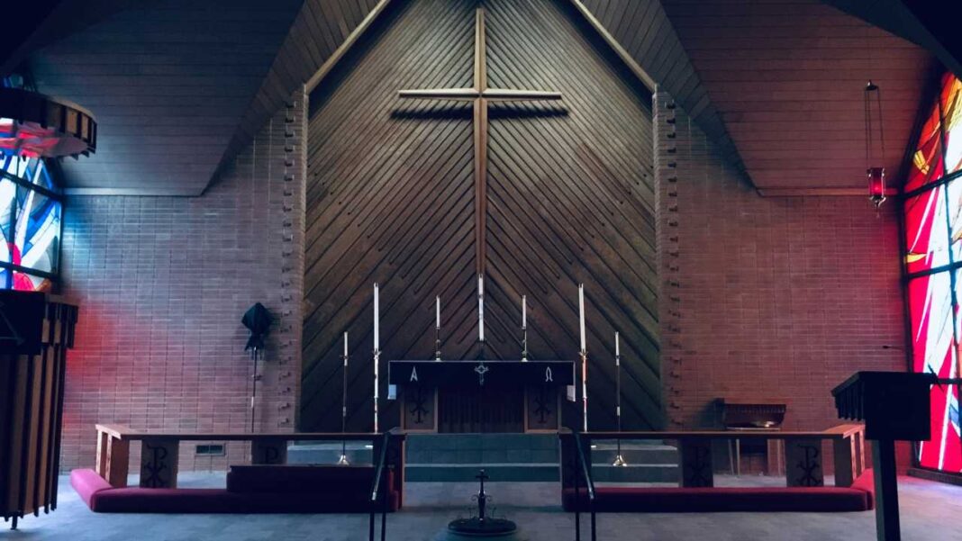 Vista frontal del interior de una iglesia con una gran cruz de madera sobre un altar, candelabros, bancos y vitrales de colores a ambos lados.