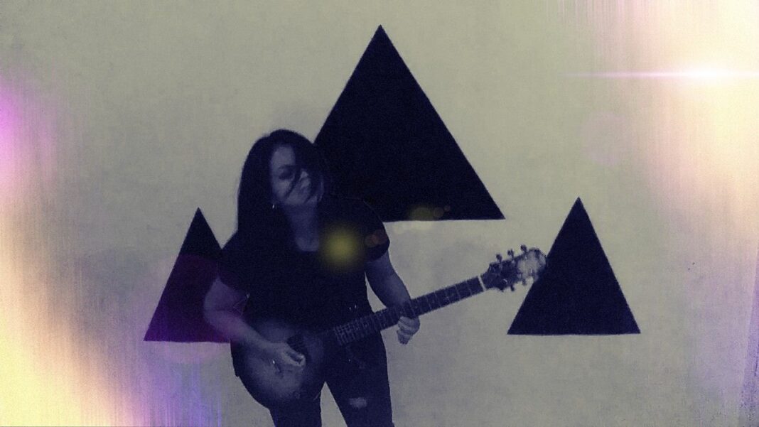 Persona de cabello oscuro sostiene una guitarra eléctrica delante de una pared clara con tres grandes triángulos negros y reflejos de luz.