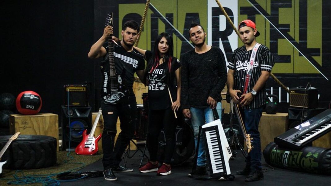 Cuatro personas posan en un estudio de música con guitarras, teclados y equipo de sonido sobre un fondo con letras grandes.