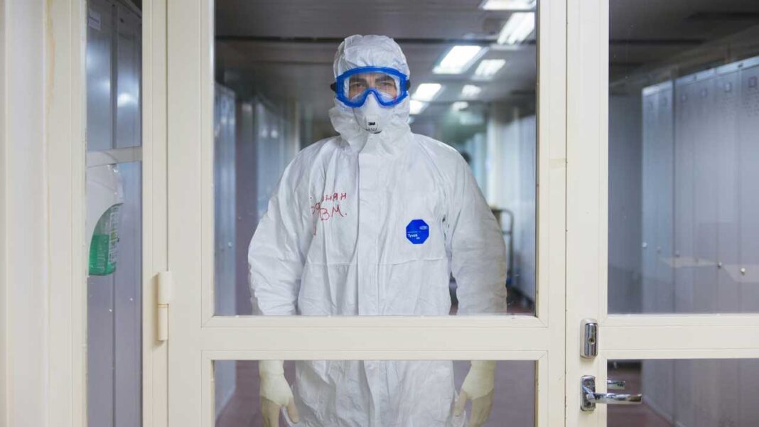 Persona con traje protector blanco, gafas azules y guantes está de pie detrás de una puerta de vidrio en un pasillo.