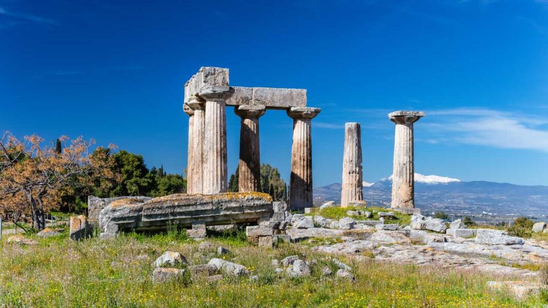 Ruinas de columnas de piedra clásicas en un prado verde, con bloques dispersos, árboles y montañas al fondo bajo un cielo azul.