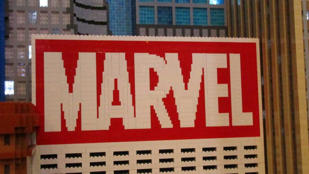 Letrero grande rojo con las letras blancas MARVEL formadas por bloques, montado sobre una fachada con ventanas y edificios al fondo.