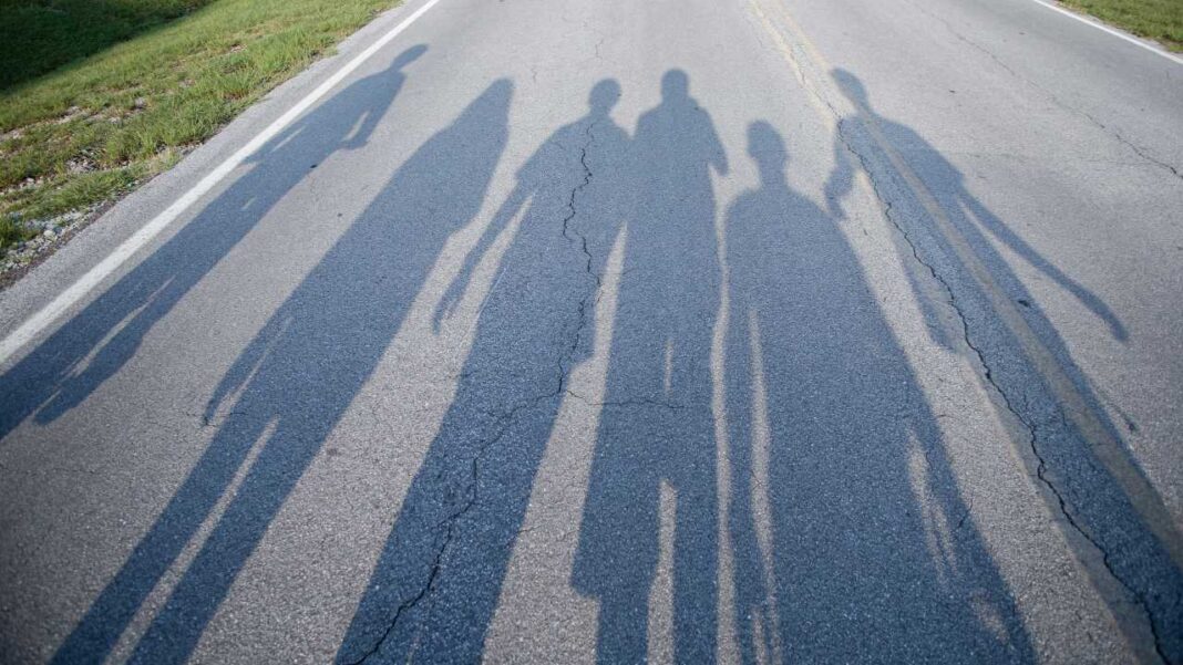 Sombras alargadas de varias personas proyectadas sobre una carretera asfaltada con una línea blanca y hierba junto al borde.