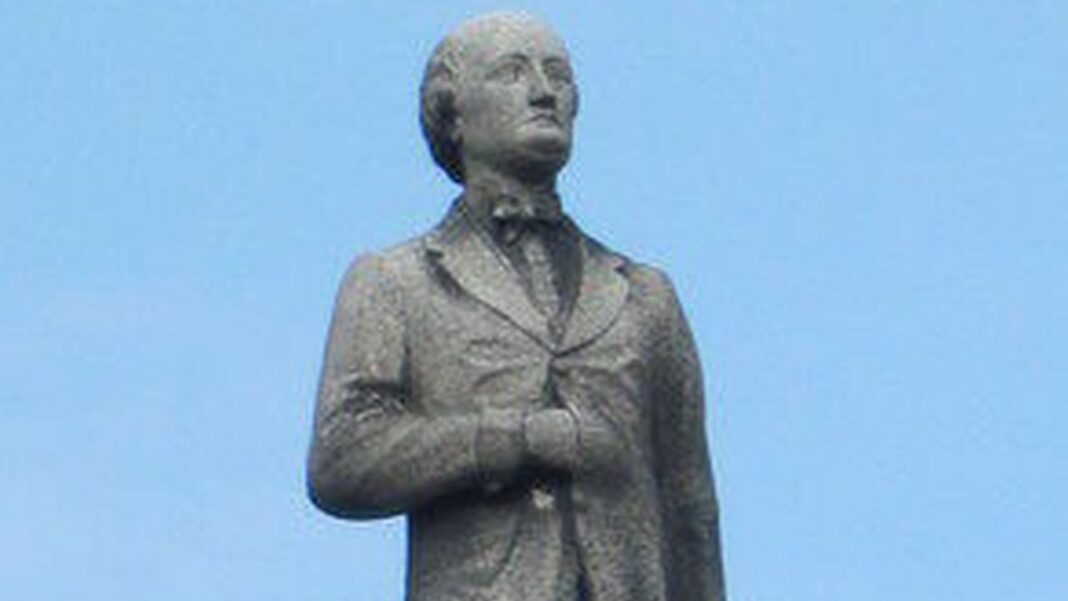 Estatua de piedra de un hombre vestido con traje y pajarita, vista de medio cuerpo contra un cielo azul despejado.