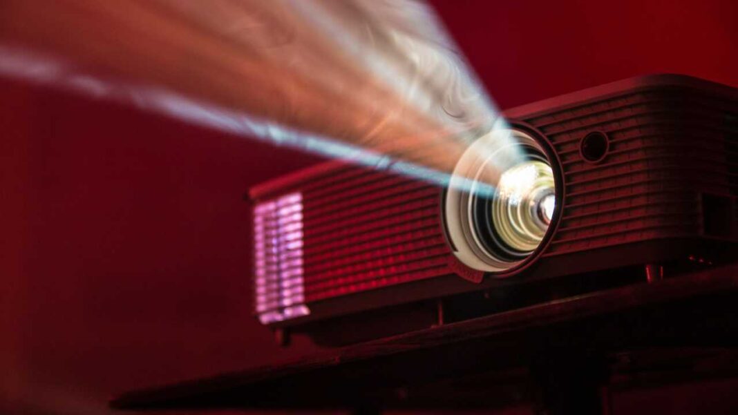 Proyector encendido sobre una superficie oscura, con la lente iluminada y un haz de luz que se extiende hacia la izquierda sobre un fondo rojo.
