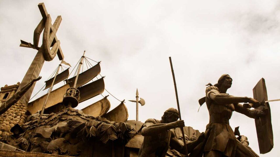 Monumento de tono bronce con figuras humanas armadas, un barco de velas y una estructura alta en forma de cruz bajo un cielo nublado.