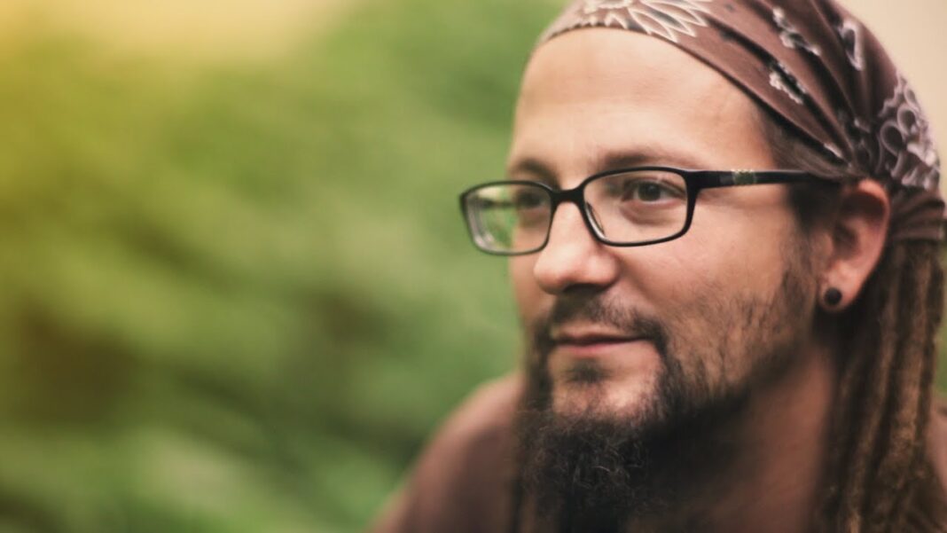 Primer plano de un hombre con gafas, pañuelo en la cabeza, rastas y barba, mirando hacia un lado sobre un fondo verde desenfocado.