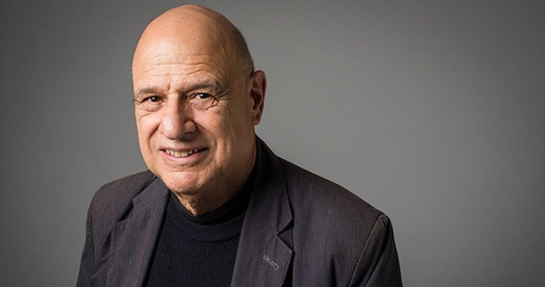 Retrato de un hombre mayor calvo y sonriente, con chaqueta oscura sobre suéter negro, frente a un fondo gris liso.