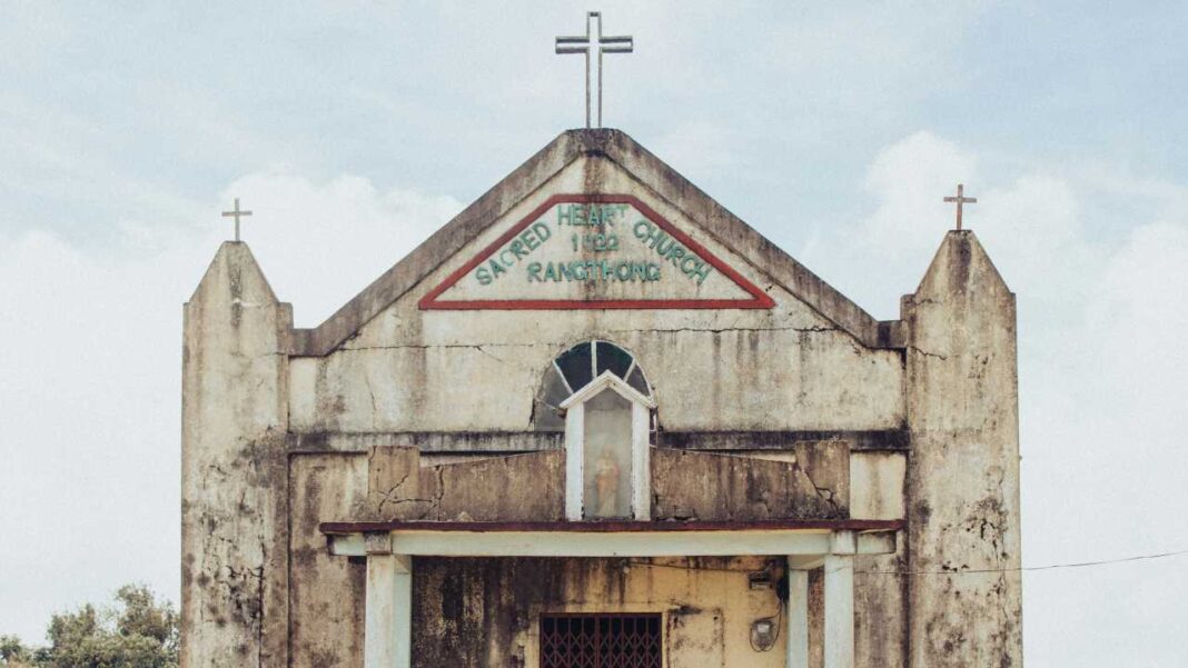 Fachada antigua de iglesia de concreto con tres cruces, letrero pintado, ventana arqueada y entrada con reja bajo cielo nublado.