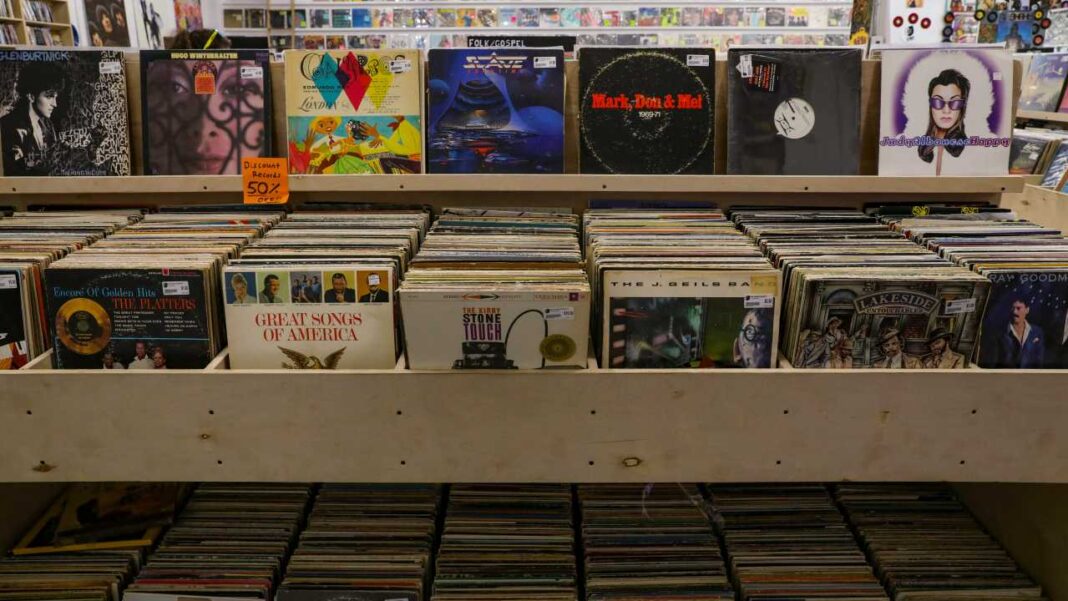Estantería de madera con filas de discos de vinilo y portadas expuestas en una tienda, con varias cubiertas de álbumes visibles y cajas inferiores llenas de discos.