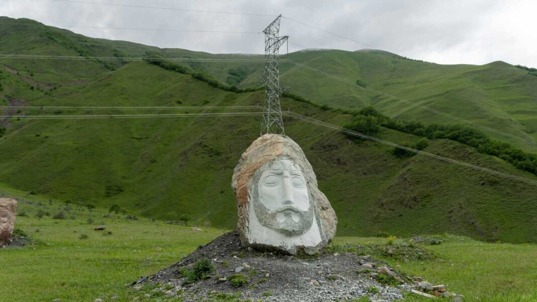 Escultura de piedra de un rostro con barba y ojos cerrados sobre un montículo de grava, ante colinas verdes, una torre eléctrica y un cielo nublado.