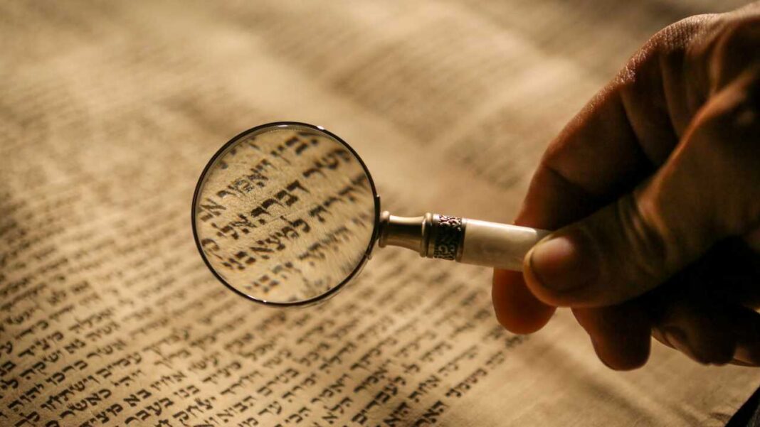 Primer plano de una mano sosteniendo una lupa sobre un manuscrito antiguo con texto hebreo, con luz cálida y fondo desenfocado.