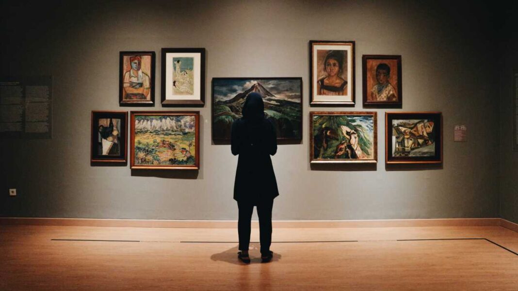 Persona de espaldas con abrigo oscuro observa una pared con varias pinturas enmarcadas en una galería iluminada, sobre un suelo de madera.