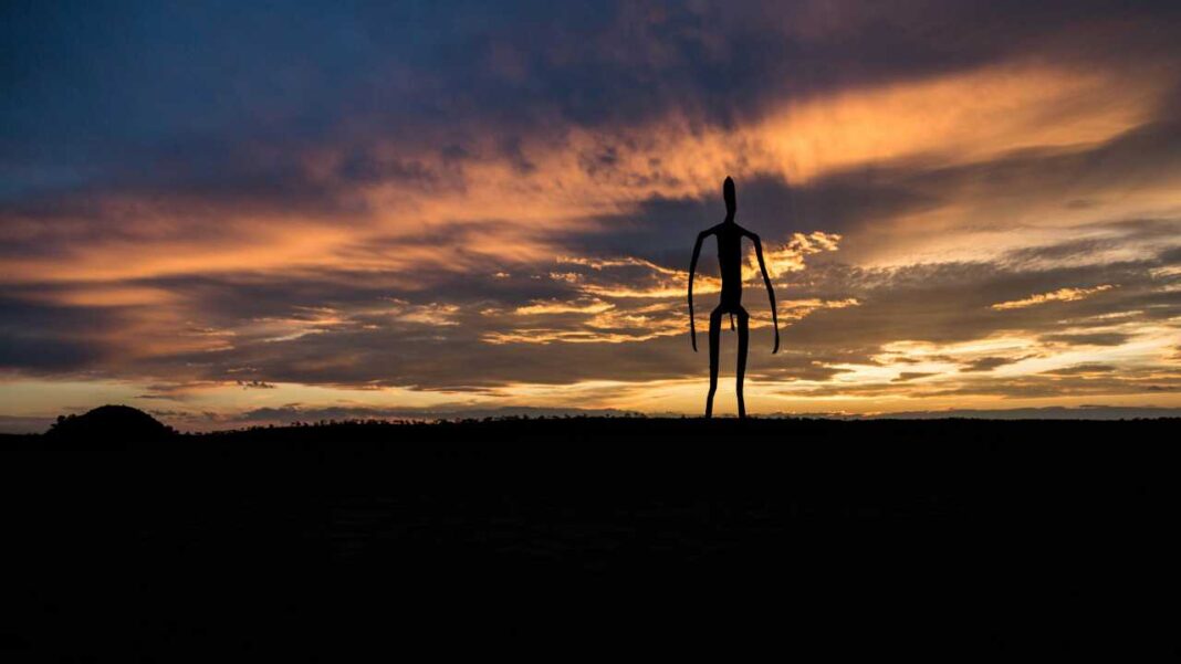 Silueta oscura de una figura humana alargada y delgada sobre un horizonte negro, bajo un cielo con nubes naranjas y azules al atardecer.