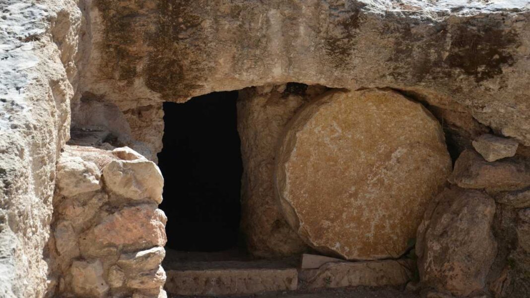 Entrada rectangular oscura en una estructura de piedra rústica, con un gran disco circular de piedra apoyado a la derecha y escalones en la base.
