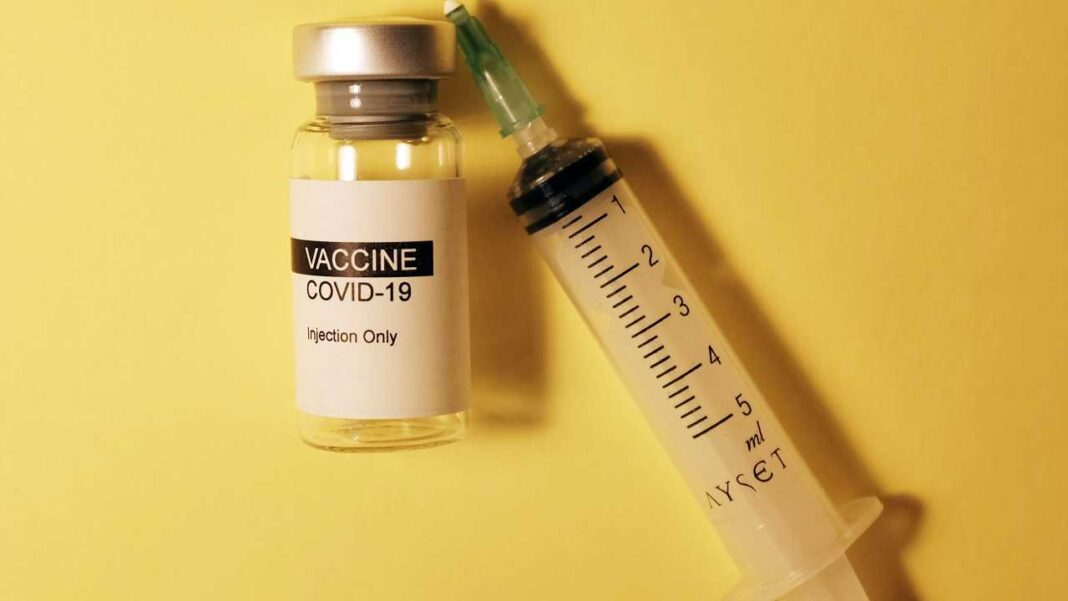 Un frasco de vidrio con etiqueta que dice VACCINE COVID-19 e Injection Only junto a una jeringa graduada con aguja verde sobre fondo amarillo.