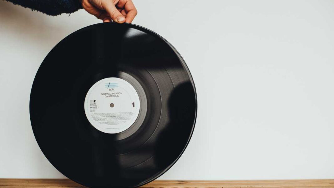 Una mano sostiene un disco de vinilo negro con etiqueta blanca que muestra el nombre Michael Jackson y Dangerous, frente a una pared blanca y una superficie de madera.