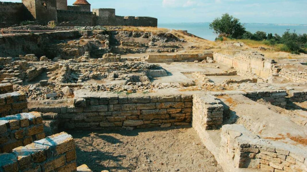 Vista de ruinas arqueológicas con muros bajos de piedra, terreno excavado, colinas secas, árboles y una masa de agua al fondo bajo un cielo claro.