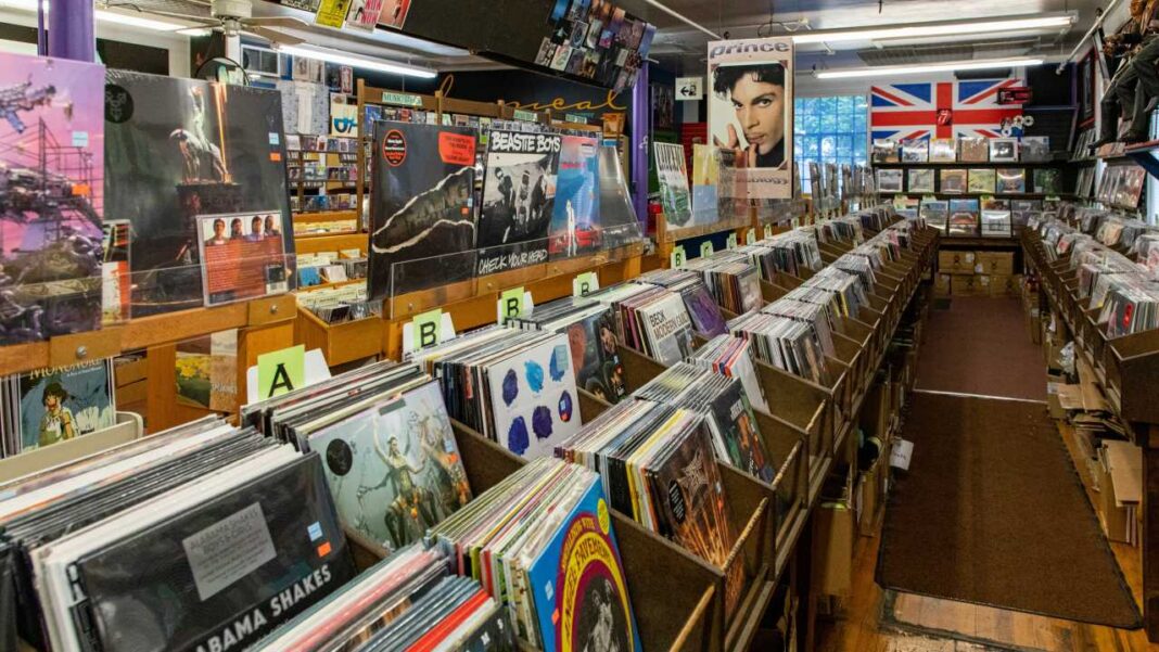 Interior de una tienda de discos con cestas de vinilos ordenados por letras, estanterías repletas, carteles musicales y una bandera británica al fondo.