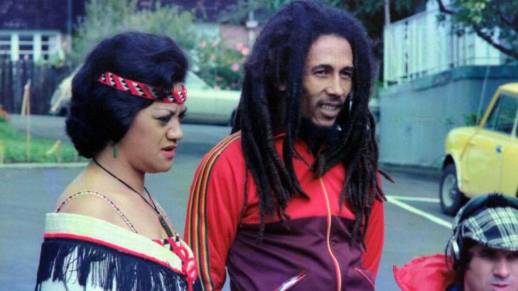 Una mujer con cinta roja en el pelo y un hombre con rastas largas y chaqueta roja están al aire libre; al fondo se ve un coche amarillo.