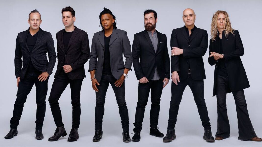 Seis hombres de pie en fila frente a un fondo gris claro, vestidos con chaquetas oscuras, camisas y pantalones negros; algunos con barba o cabello largo.