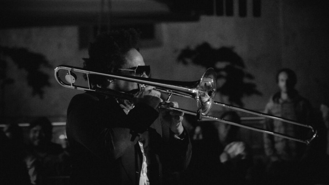 Fotografía en blanco y negro de un músico con gafas de sol tocando un trombón ante un público desenfocado en un espacio interior oscuro.