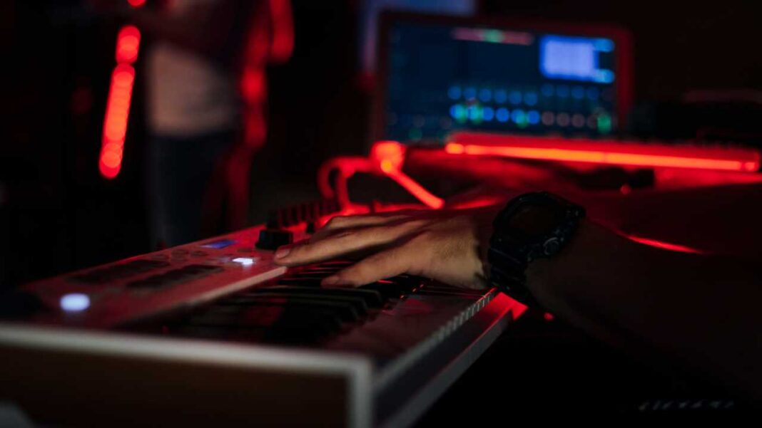 Primer plano de una mano con reloj sobre un teclado musical, con luces rojas y una pantalla de computadora desenfocada al fondo.