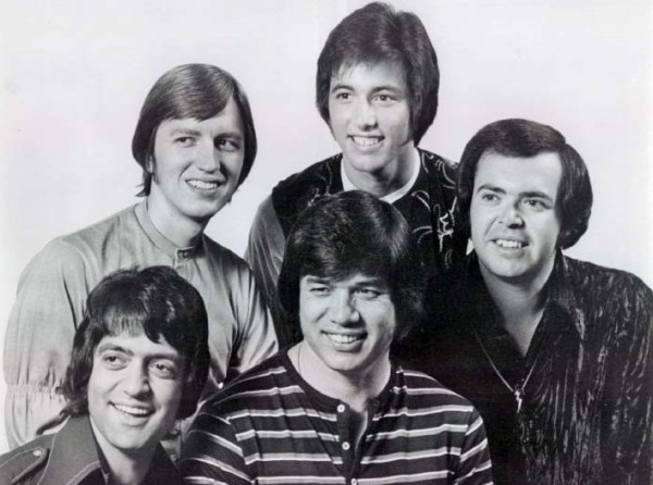 Retrato en blanco y negro de cinco hombres jóvenes sonriendo, con peinados de los años setenta y camisas de distintos estilos.