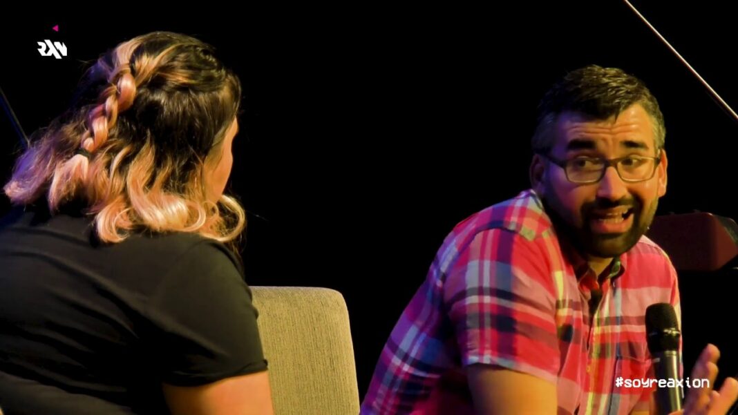 Dos personas en un escenario oscuro: una mujer de espaldas con cabello trenzado y un hombre con gafas y camisa a cuadros hablando con un micrófono.