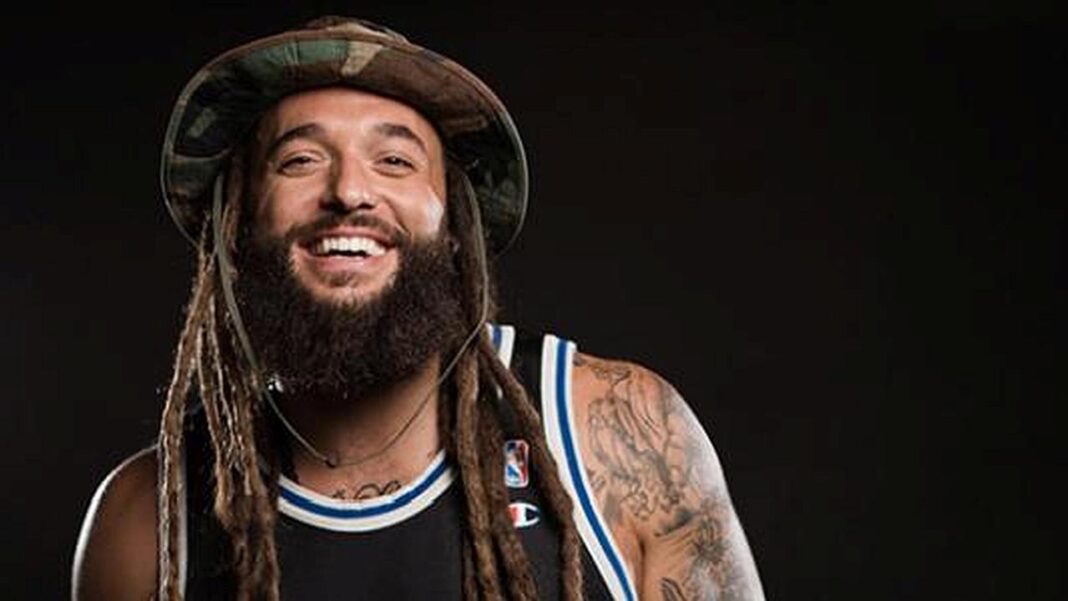 Retrato de un hombre sonriente con barba, rastas largas, sombrero camuflado y camiseta de baloncesto negra con ribetes azules y blancos.