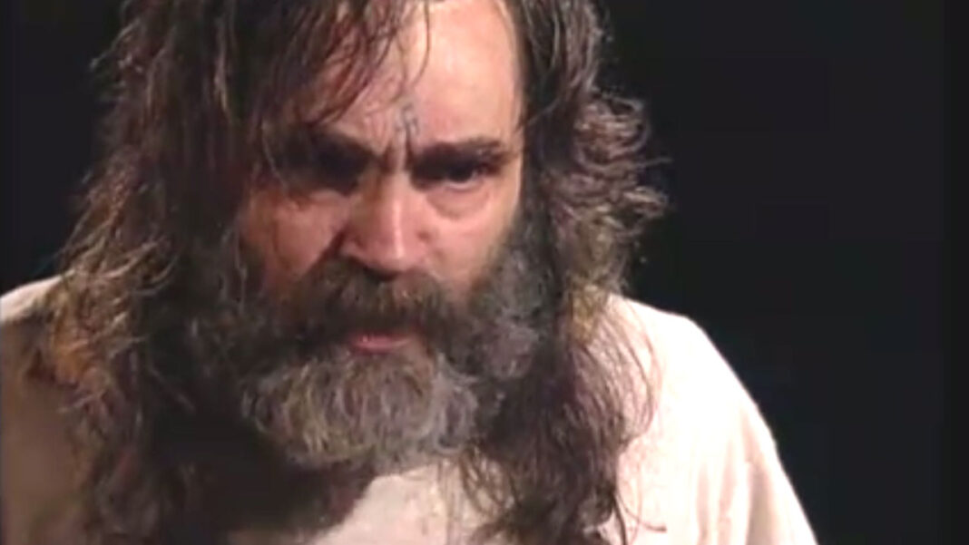 Primer plano de un hombre de pelo largo y barba espesa grisácea, con ceño fruncido y mirada baja, sobre fondo oscuro.