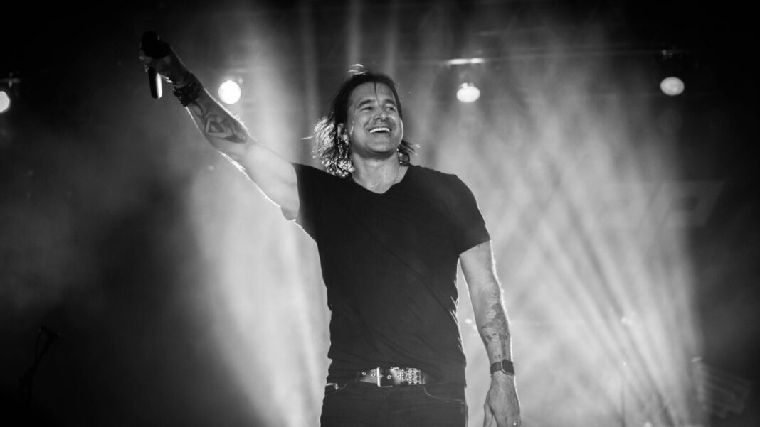 Cantante de pelo largo y camiseta negra con micrófono en alto sobre un escenario, entre haces de luz y humo en blanco y negro.