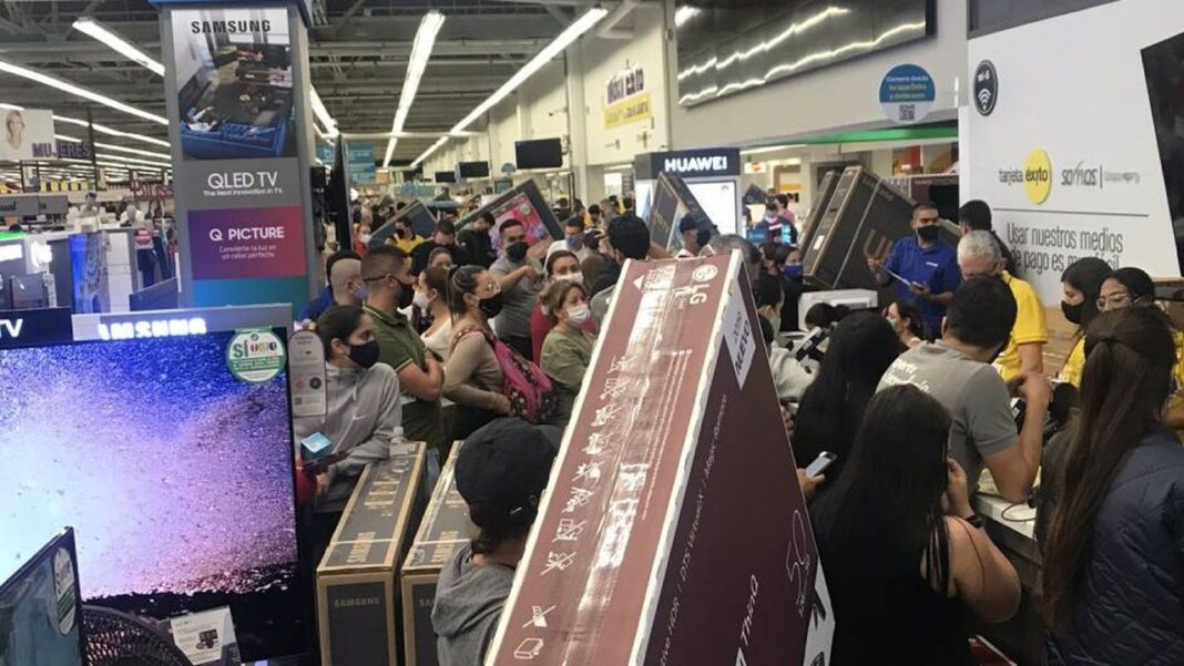 Multitud de personas con mascarillas esperando junto a cajas grandes de televisores en una tienda de electrónica con letreros de Samsung y Huawei.