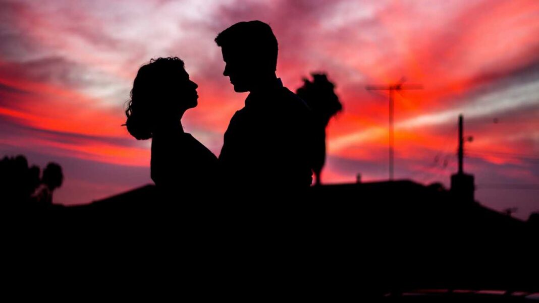 Silueta de una pareja de perfil frente a un cielo rojo y nubes intensas, con tejados y una antena al fondo.