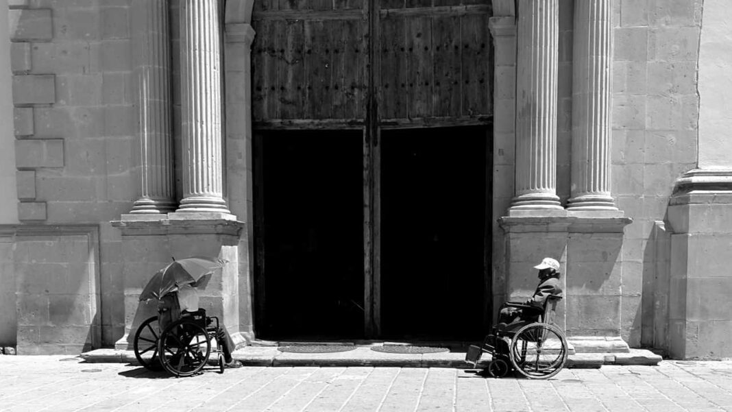 Fotografía en blanco y negro de dos personas en sillas de ruedas junto a una gran puerta de madera entre columnas de piedra.